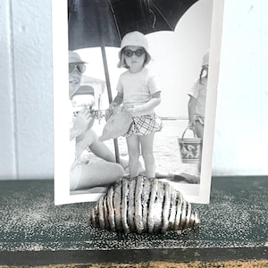 May include: A silver metal seashell shaped picture frame holding a black and white photo of a young girl wearing sunglasses and a hat at the beach.
