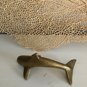 May include: A small, gold-colored brass dolphin figurine. The dolphin is facing right and has a slightly curved tail. The figurine is sitting on a white surface.