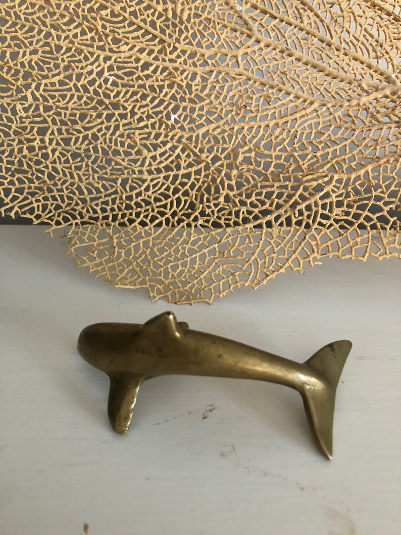 May include: A small, gold-colored brass dolphin figurine. The dolphin is facing right and has a slightly curved tail. The figurine is sitting on a white surface.
