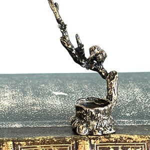 May include: A silver-toned metal sculpture of a bird perched on a branch, with the branch extending upwards and ending in a small, rounded stump. The sculpture is detailed with textured bark and leaves.