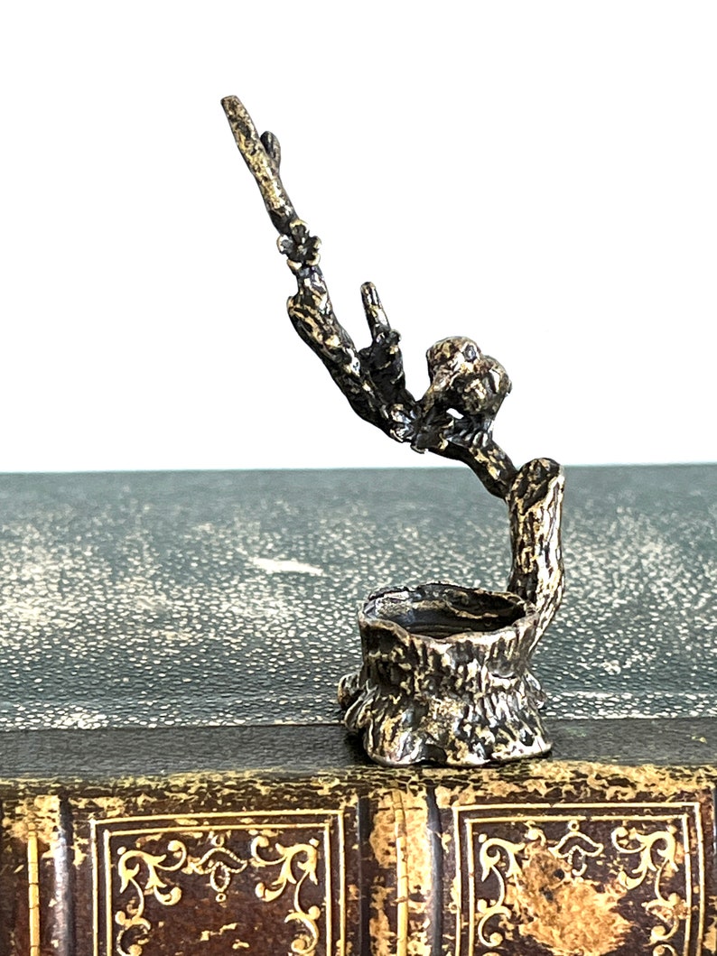 May include: A silver-toned metal sculpture of a bird perched on a branch, with the branch extending upwards and ending in a small, rounded stump. The sculpture is detailed with textured bark and leaves.