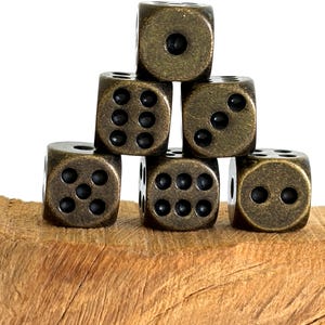 May include: A stack of six metallic dice with an antique bronze finish. The dice are arranged in a pyramid formation, displaying various black-pitted numerical faces including one, two, three, five, and six, with rounded corners and edges.