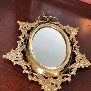 Puede incluir: Un espejo ovalado dorado con un marco decorativo y ornamentado. El marco tiene un dise&ntilde;o floral y est&aacute; unido a un anillo para colgar.