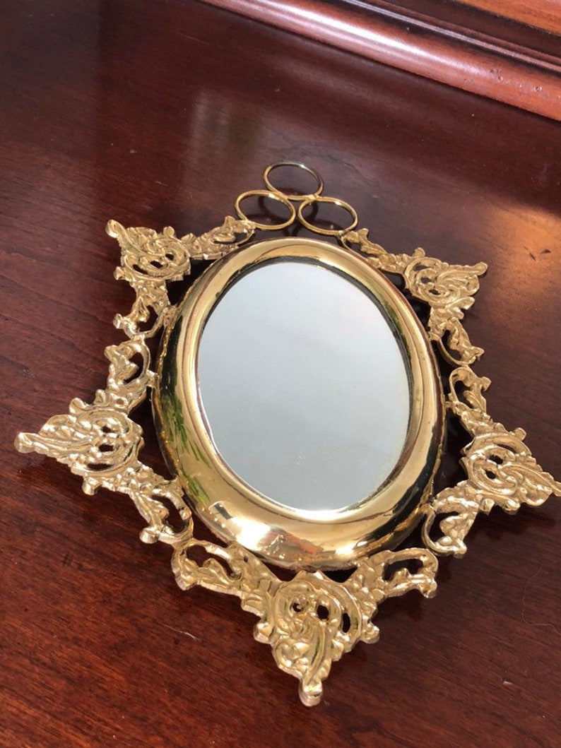 Puede incluir: Un espejo ovalado dorado con un marco decorativo y ornamentado. El marco tiene un dise&ntilde;o floral y est&aacute; unido a un anillo para colgar.