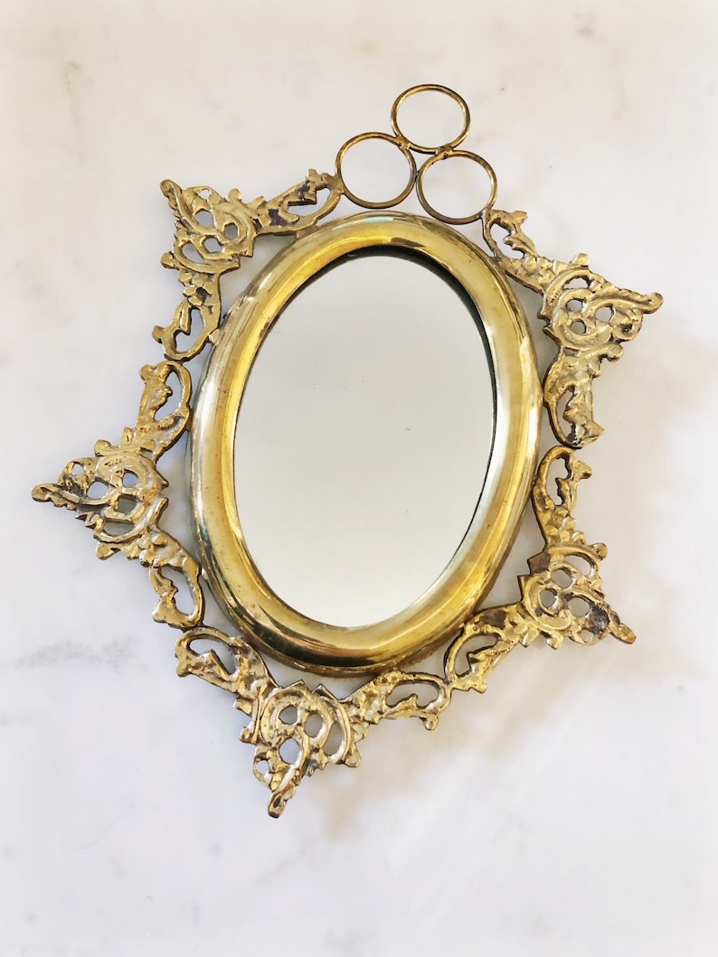 Puede incluir: Un espejo ovalado dorado con un marco ornamentado y decorativo. El marco tiene un dise&ntilde;o floral delicado y un colgador en forma de anillo en la parte superior.