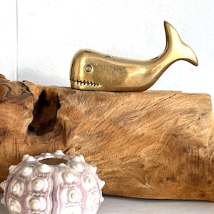May include: A brass whale figurine with a detailed tail and mouth rests on a piece of wood. A sea urchin shell with a white and purple pattern sits below the whale. The wood has a natural, textured appearance.