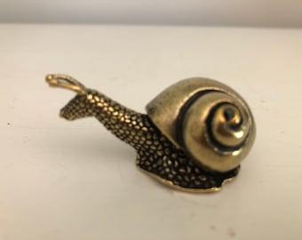 May include: A small brass snail figurine with a textured, bumpy surface on its body and a smooth, spiraled shell. The metal has a muted gold tone with darker, aged patina accents in the crevices of the shell and along the neck.