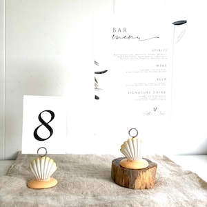 May include: A white table number card with the black number 8, a white bar menu card with black text, and two seashell-shaped card holders with metal wire clips. One holder sits directly on the surface, while the other rests on a small wood slice.