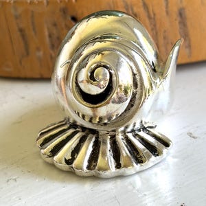 May include: A silver-toned metal figurine shaped like a snail with a prominent spiral shell pattern and a wide, fluted base. The object features a polished, reflective surface with dark recessed areas that highlight the intricate, swirling shell design.