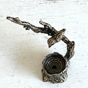 May include: A bronze sculpture of a bird perched on a branch with a nest at the base. The sculpture is detailed with textured bark and a spiral-shaped nest.