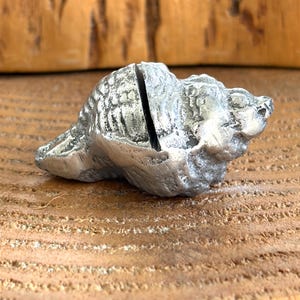 May include: A silver-toned metal seashell figurine featuring a textured, weathered surface and a distinct vertical slit cut into the center of the shell. The object has a metallic finish with dark gray recessed areas that highlight the natural ridges.