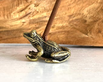 May include: A small, bronze-colored frog incense holder with a dark brown incense stick. The frog is detailed with textured skin and sits on a white surface. The background features a wooden texture.