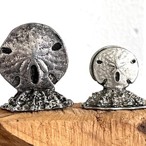 May include: Two silver-toned sand dollar sculptures. The larger one is upright, while the smaller one is to the right. Both have detailed textures and are mounted on a textured base, evoking a coastal theme.