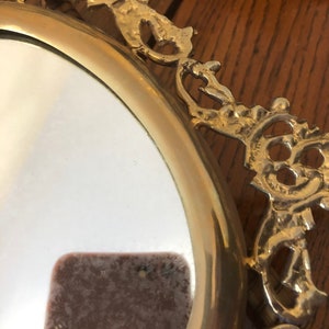 Puede incluir: Un espejo ovalado con un marco decorativo y ornamentado. El marco est&aacute; hecho de metal dorado y tiene un dise&ntilde;o detallado e intrincado.