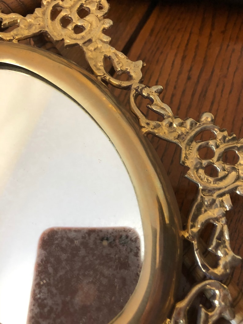 Puede incluir: Un espejo ovalado con un marco decorativo y ornamentado. El marco est&aacute; hecho de metal dorado y tiene un dise&ntilde;o detallado e intrincado.