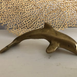 May include: A small, solid brass dolphin figurine. The dolphin is in a leaping pose with its tail raised and its head tilted down.
