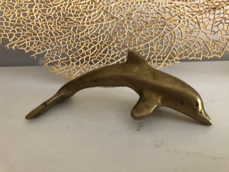 May include: A small, solid brass dolphin figurine. The dolphin is in a leaping pose with its tail raised and its head tilted down.