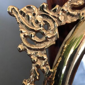 Puede incluir: Primer plano de un marco de espejo ovalado adornado con intrincados detalles. El marco est&aacute; hecho de metal dorado y tiene un acabado altamente pulido.