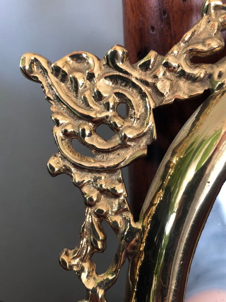 Puede incluir: Primer plano de un marco de espejo ovalado adornado con intrincados detalles. El marco est&aacute; hecho de metal dorado y tiene un acabado altamente pulido.