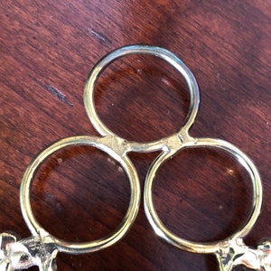 Puede incluir: Un soporte decorativo de metal dorado con tres anillos circulares y detalles ornamentados. El soporte tiene una parte superior redonda y est&aacute; dise&ntilde;ado para sostener una variedad de art&iacute;culos.