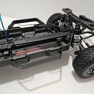 May include: A black and gray remote control truck chassis with large, knobby tires. The chassis features a blue component and a black roll cage. The suspension includes white coil springs. The truck is designed for off-road use.