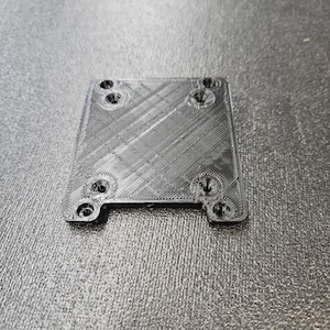May include: A black plastic square plate with six holes for screws. The plate has a textured surface.