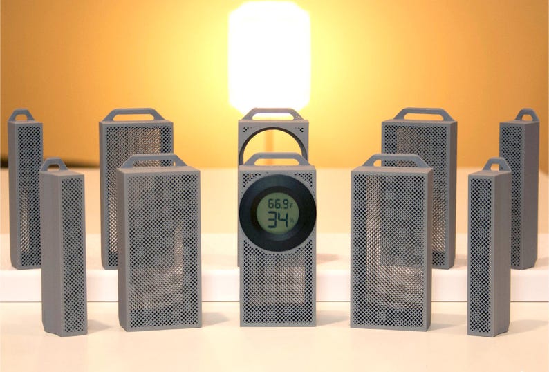 May include: A collection of gray, rectangular, perforated speakers with handles. One speaker has a digital display showing temperature and humidity readings. The speakers are arranged on a white surface against a warm, yellow background.