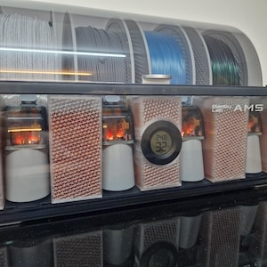 May include: A black and clear 3D printer filament storage unit with the text "Bambu Lab AMS 2". The unit has multiple compartments for filament spools and internal components. A digital display shows temperature readings.