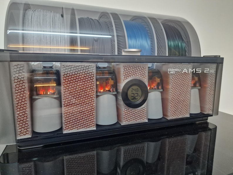 May include: A black and clear 3D printer filament storage unit with the text "Bambu Lab AMS 2". The unit has multiple compartments for filament spools and internal components. A digital display shows temperature readings.