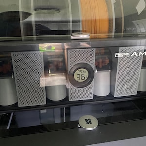 May include: A black and gray Bambu Lab AMS 2 unit with a clear top, displaying multiple filament spools. The unit has a digital display showing temperature readings. The front panel features a perforated design.