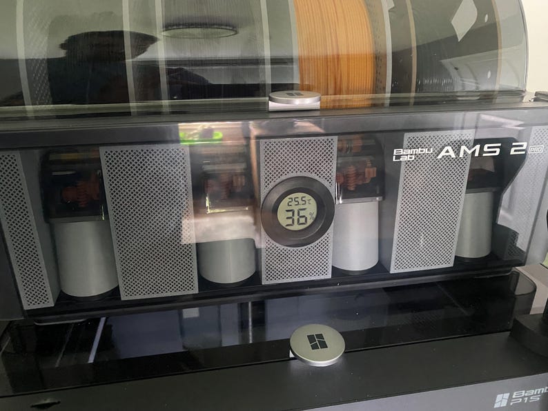 May include: A black and gray Bambu Lab AMS 2 unit with a clear top, displaying multiple filament spools. The unit has a digital display showing temperature readings. The front panel features a perforated design.