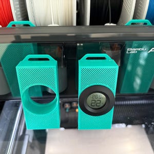 May include: Two teal 3D-printed holders with a perforated design. One has a circular opening, the other has a digital display showing 26.9 and 22. The holders are in front of a black and teal device with the text "Bambu Lab AMS".