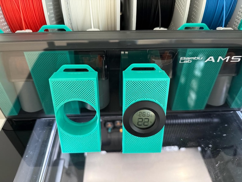 May include: Two teal 3D-printed holders with a perforated design. One has a circular opening, the other has a digital display showing 26.9 and 22. The holders are in front of a black and teal device with the text "Bambu Lab AMS".