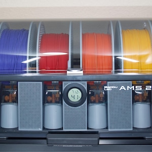 May include: A black and gray Bambu Lab AMS 2 printer with four spools of filament in blue, red, orange, and yellow. The printer has a digital display showing the temperature. The printer is designed for 3D printing.