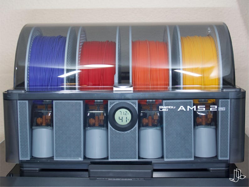 May include: A black and gray Bambu Lab AMS 2 printer with four spools of filament in blue, red, orange, and yellow. The printer has a digital display showing the temperature. The printer is designed for 3D printing.