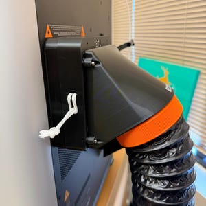 May include: A black plastic exhaust adapter attached to the back of a machine, featuring a white cord loop on the side, a bright orange textured ring, and a flexible black accordion-style ventilation hose extending downward from the unit.