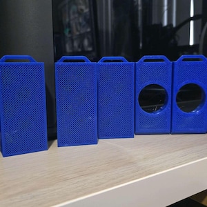 May include: A collection of blue 3D-printed rectangular cases with a textured surface. Some cases have a handle at the top, while others have circular cutouts. The objects are arranged on a white shelf.