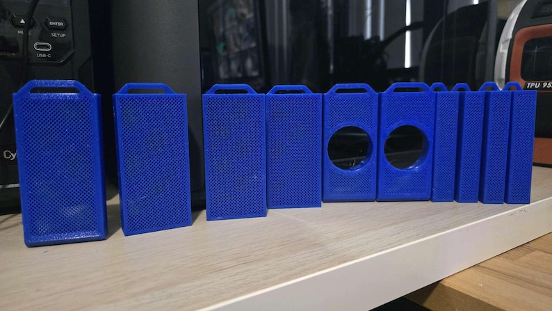 May include: A collection of blue 3D-printed rectangular cases with a textured surface. Some cases have a handle at the top, while others have circular cutouts. The objects are arranged on a white shelf.
