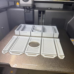 May include: A 3D printer with several light gray, rectangular, and circular components. The components have a perforated design and are arranged on the printer's surface. The printer has a black and gray body with the brand name "Bambu Lab".