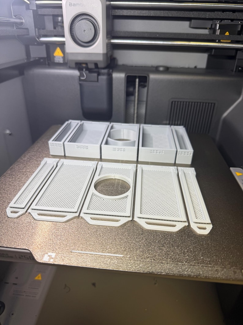 May include: A 3D printer with several light gray, rectangular, and circular components. The components have a perforated design and are arranged on the printer's surface. The printer has a black and gray body with the brand name "Bambu Lab".