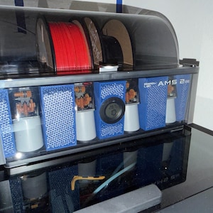 May include: A 3D printer filament storage and dispensing unit. The unit has a clear, tinted top and a silver and blue body. It contains red filament spools, and has a digital temperature display. The text "AMS 2" is visible.