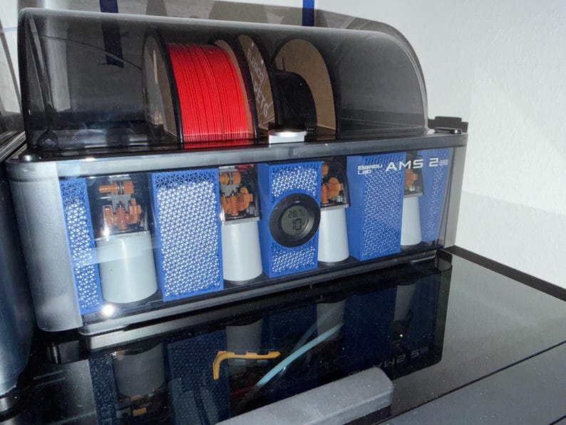 May include: A 3D printer filament storage and dispensing unit. The unit has a clear, tinted top and a silver and blue body. It contains red filament spools, and has a digital temperature display. The text "AMS 2" is visible.