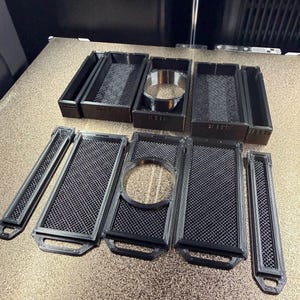 May include: A collection of black 3D-printed parts, including rectangular trays with mesh inserts and circular components. The parts are arranged on a light-colored surface, likely for a 3D printer.