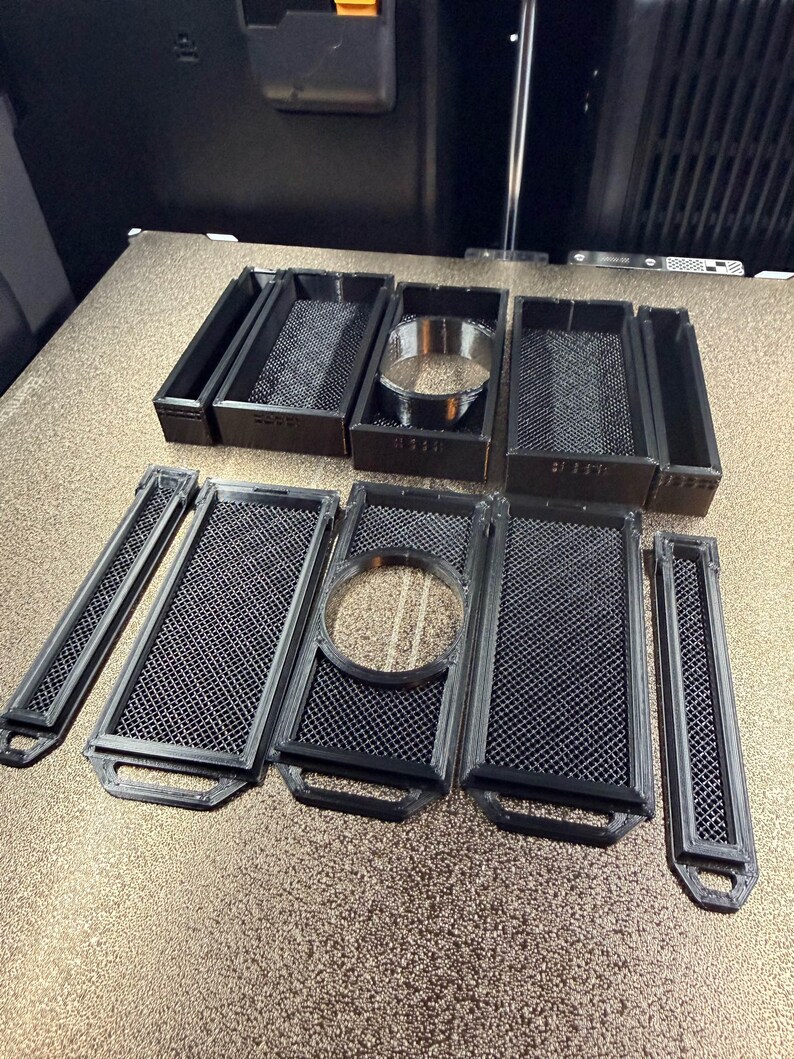 May include: A collection of black 3D-printed parts, including rectangular trays with mesh inserts and circular components. The parts are arranged on a light-colored surface, likely for a 3D printer.