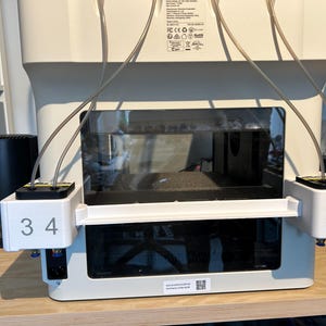 May include: A white 3D printer featuring a multi-material system attachment on the front. The attachment has two white blocks labeled with the numbers 3, 4 on the left and 2, 1 on the right, with thin grey tubes connecting to the top of the printer.
