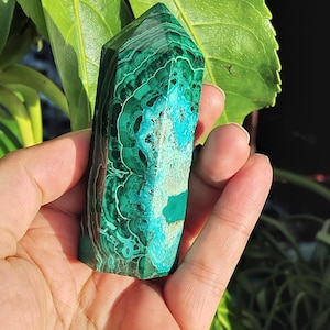 May include: A polished malachite crystal tower featuring deep forest green concentric banding patterns and a vibrant turquoise blue mineral inclusion. The stone is shaped into a hexagonal point with a smooth, reflective surface and natural textures.