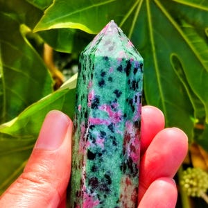 May include: A green and pink ruby zoisite crystal tower. The polished stone has a rectangular shape with a pointed top. The crystal is held in front of a background of green leaves.
