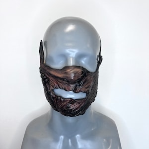 以下が含まれることがあります： 茶色と黒のマスクで、質感のある有機的なデザインで、様式化されたあごひげと口に似ています。 マスクは灰色のマネキンの頭に着用され、固定するための黒いストラップが付いています。 マスクのディテールには、暗く風化した外観が含まれています。