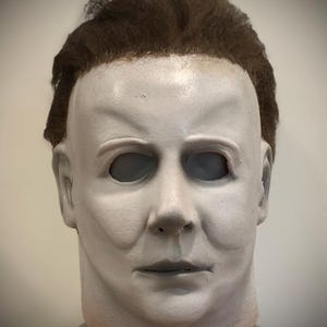 Pode incluir: Uma máscara de Halloween branca com uma peruca castanha. A máscara tem um rosto realista com uma expressão neutra.