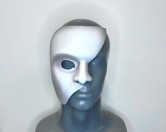 May include: A white half mask covering one half of a mannequin's face. The mask is made of a smooth, white material and has a simple design. The mannequin's face is painted a light gray color.
